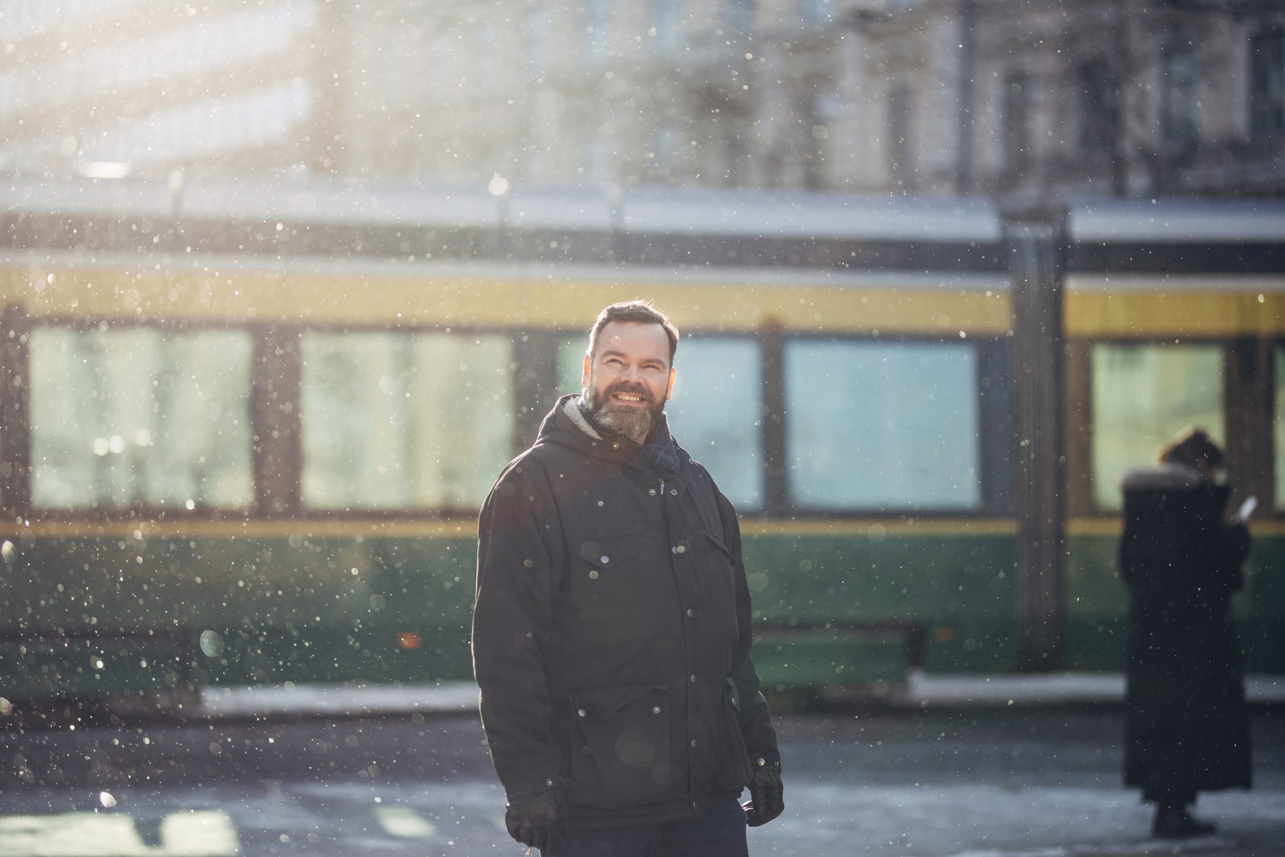 Mies seisoo lumisateessa, taustalla raitiovaunu.