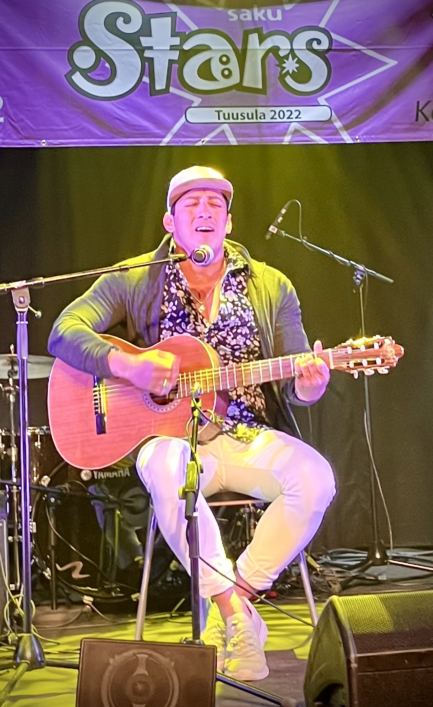 Lippalakkipäinen mies istuu lavalla soittaen kitaraa ja laulaen.