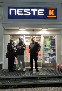 Kolme ihmistä seisoo iltavalaistuksessa Neste K -kyltin alla myymälän edessä.