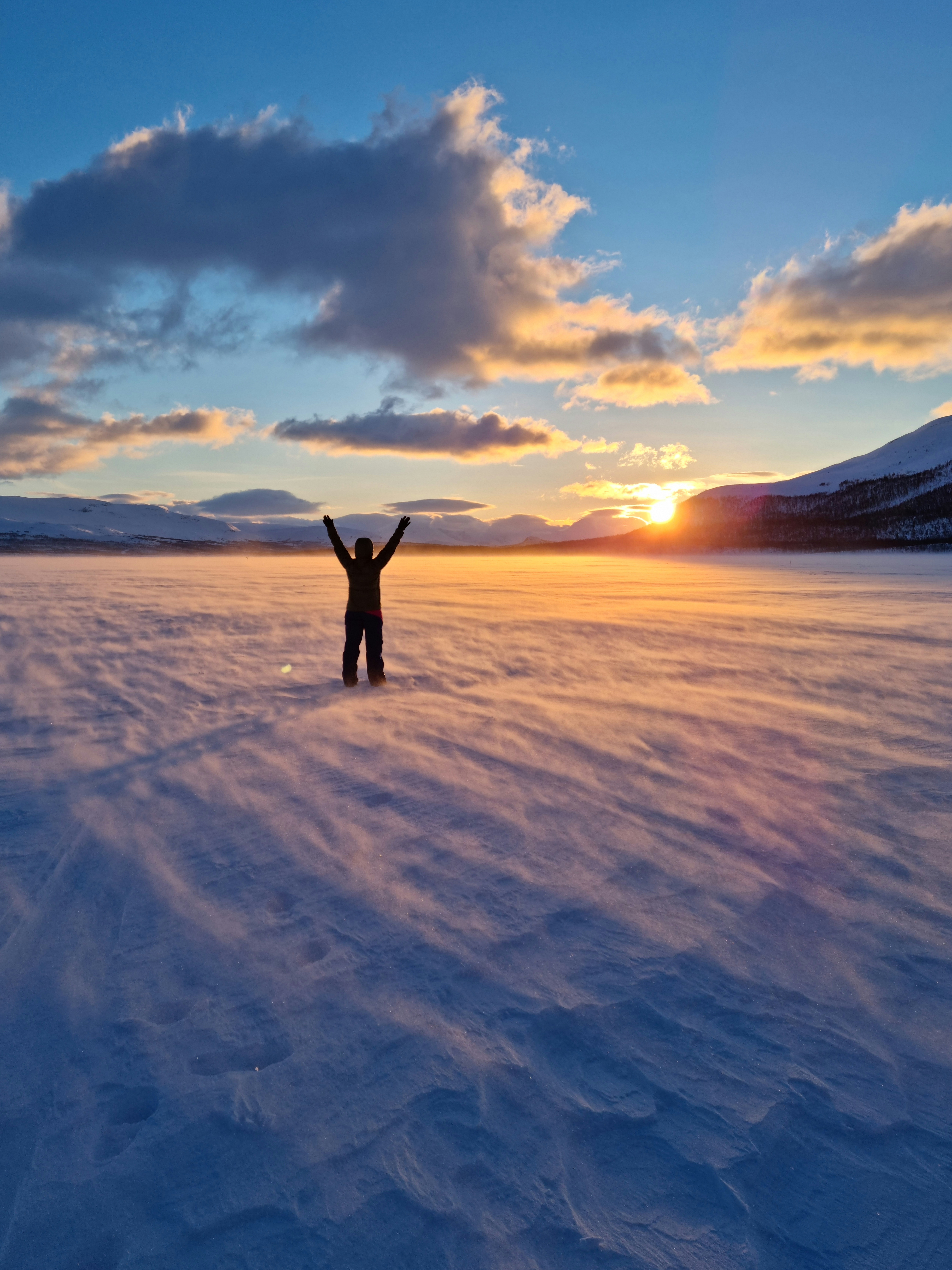 Ihminen seisoo lumisessa tunturimaisemassa kädet ylhäällä auringon laskiessa tunturien taakse.