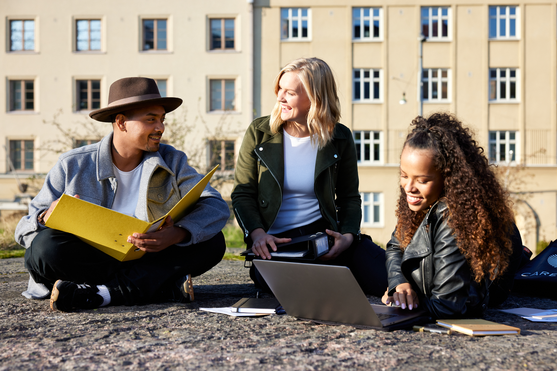 Kolme opiskelijaa istuu kalliolla mukanaan opiskelukansio ja kannettava tietokone.