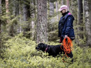 Musta koira ja lippalakkipäinen nainen metsässä.