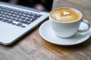 Kannettava tietokone, jonka vieressä kuppi kahvia ja pintaan piirretty vaahdolla sydän.