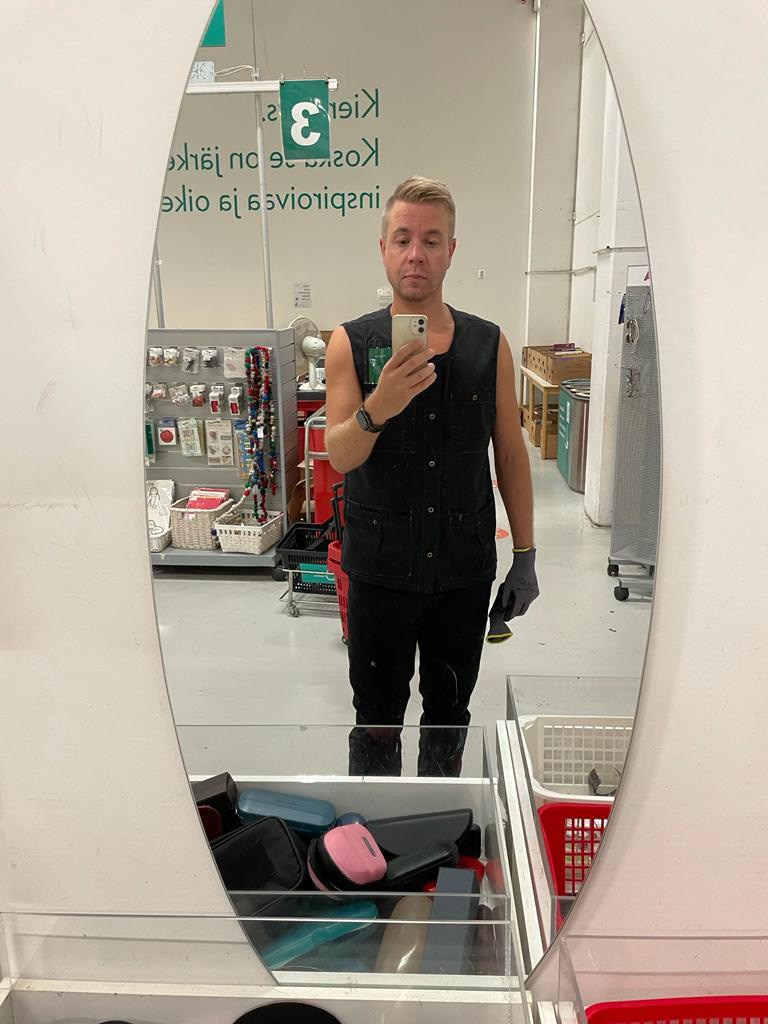 Mies seisoo kierrätysmyymälässä peilin edessä katsoen matkapuhelimeen.