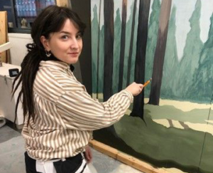 Tummahiuksinen nainen polvillaan maalaamassa metsämaisemaa.