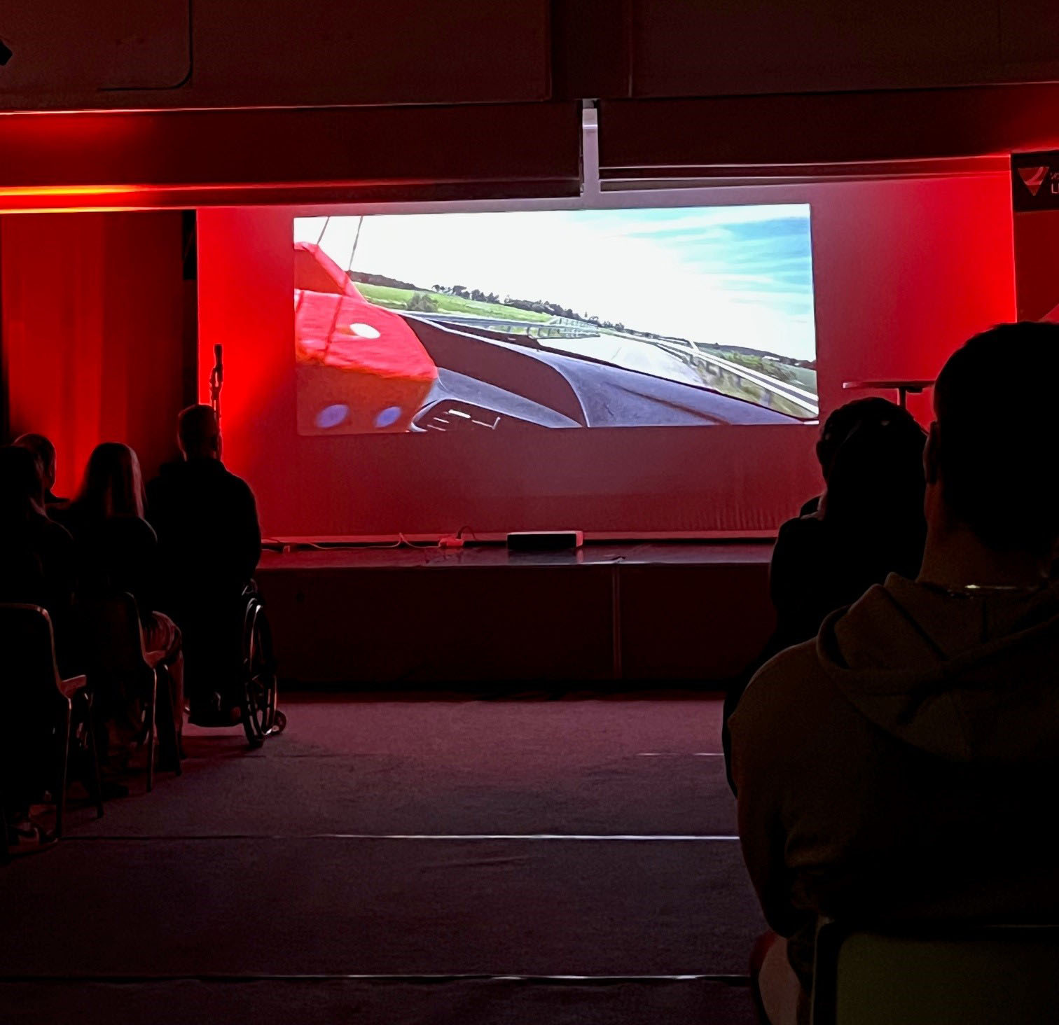 Yleisöä katsomassa Liikenneturvan videota ylinopeutta ajavasta autosta, jossa etualalla näkyy punaiset karvanopat ja edessä aukeava tie.