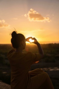 Nutturapäinen nuori nainen tekee käsillään sydämen, taustalla kullanvärinen auringonlasku.