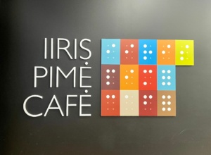 Mustalla pohjalla teksti: Iiris Pimé Café ja sama värikkäillä laatoilla pistekirjoituksella.