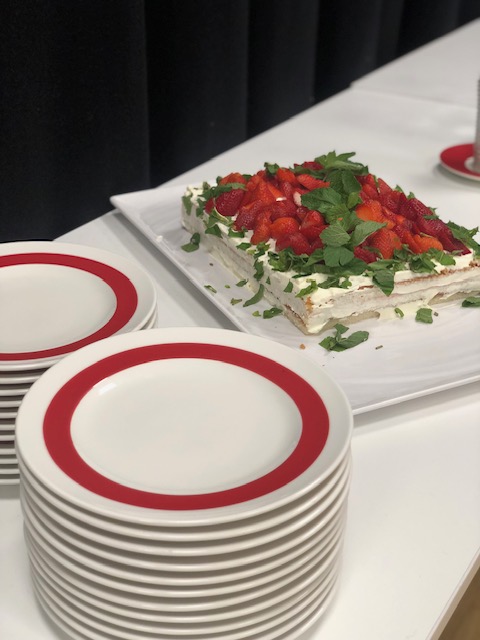 Valkoisia punaisella reunuksella varustettuja lautasia pinossa ja lautasten vieressä neliön  muotoinen mansikkakakku.