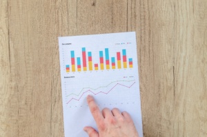 Pöydällä paperi, jossa on pylväsdiagrammi ja kehitystä kuvaava käyrä, jota ihmisen sormi osoittaa.