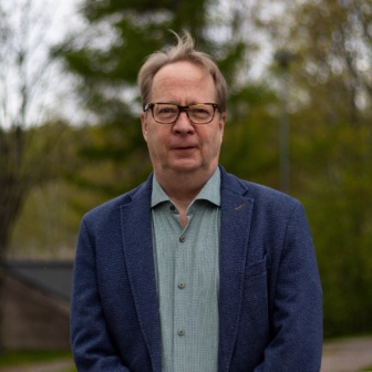 Silmälasipäinen mies pikkutakki yllään vihreää luontotaustaa vasten kuvattuna.