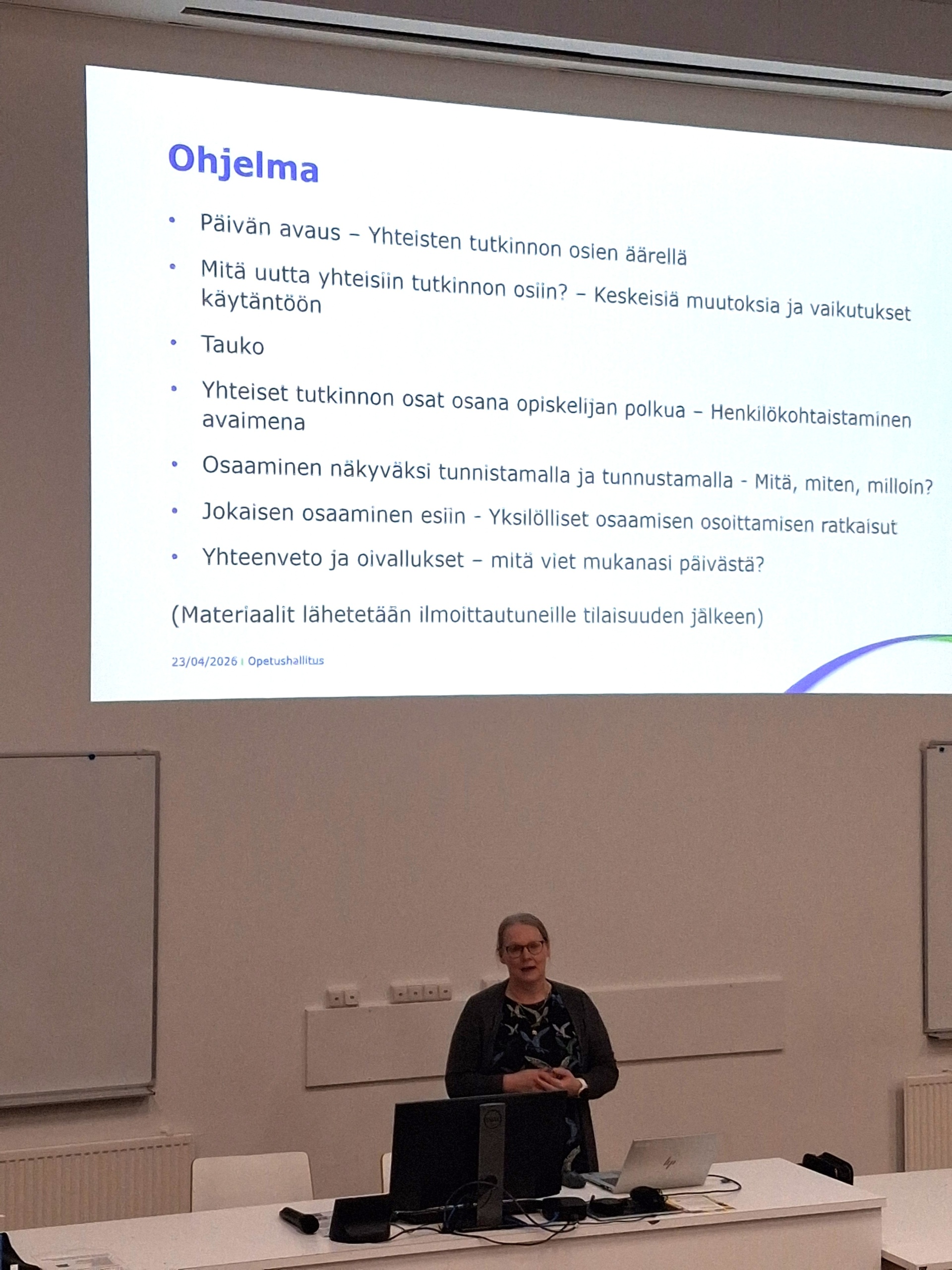 Nutturapäinen nainen pitämässä puheenvuoroa auditorion edessä. Taustalla esityksen ohjelma.