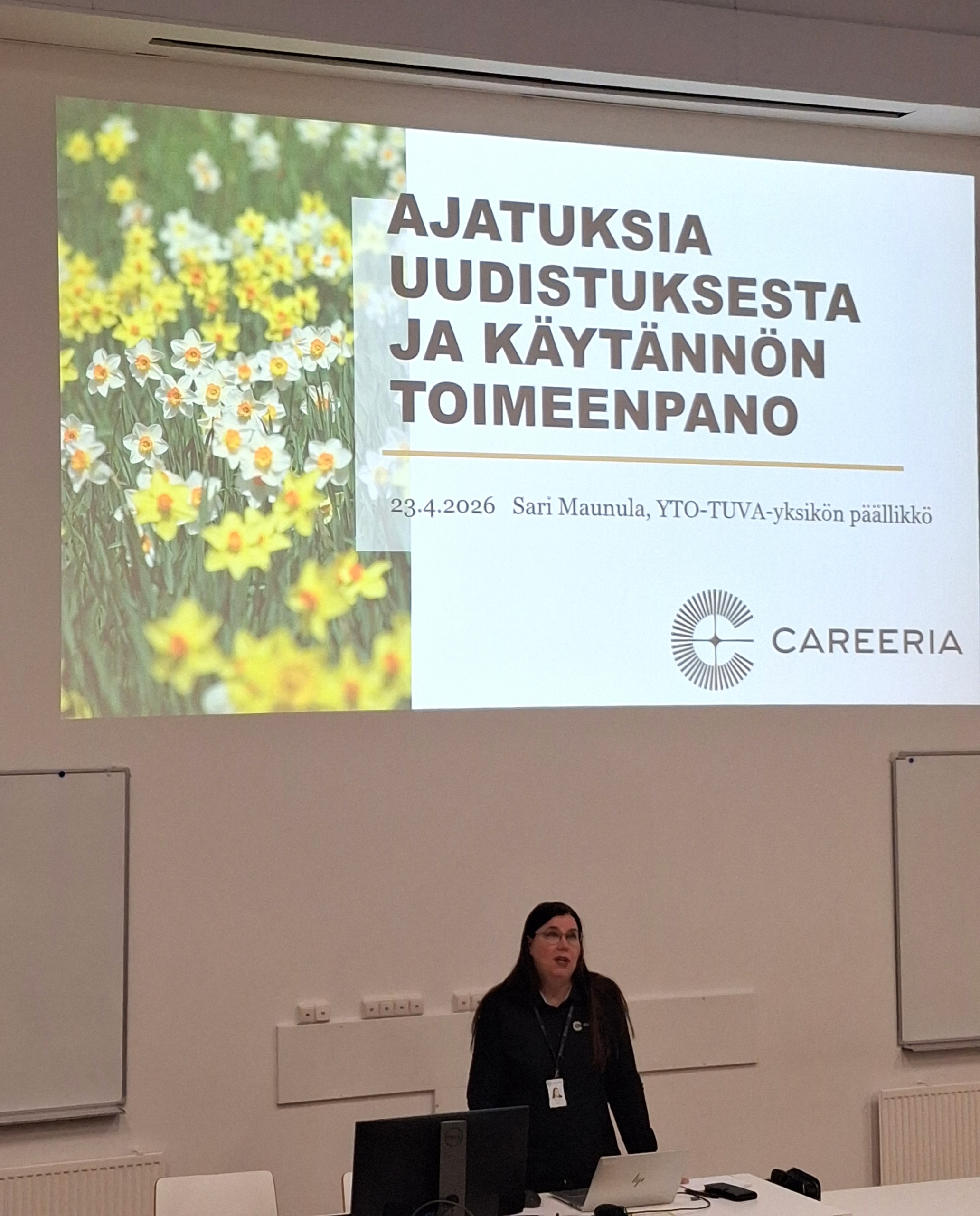 Tummahiuksinen nainen auditorion etuosassa. Taustalla kuva, jossa on hänen esityksensä otsikko: Ajatuksia uudistuksesta ja käytännön toimeenpano.