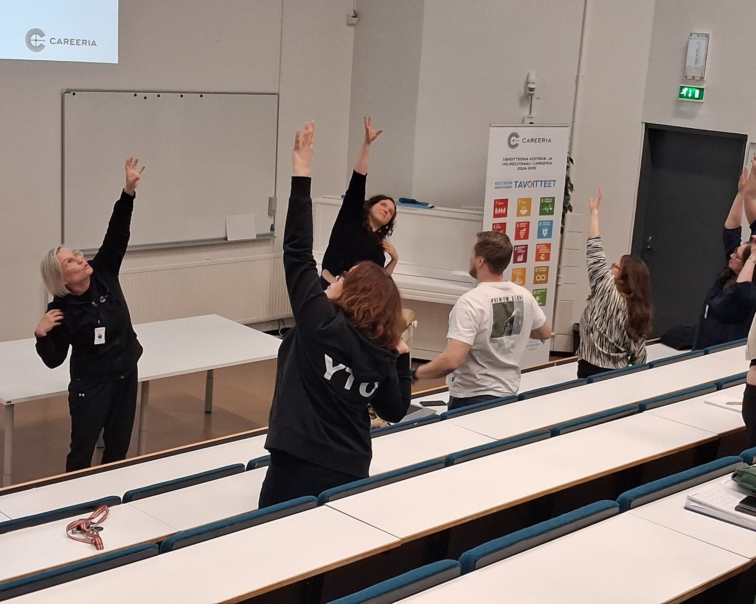 Kaksi liikunnanopettajaa auditorion edessä vetämässä taukojumppaa auditoriossa olevalle yleisölle.