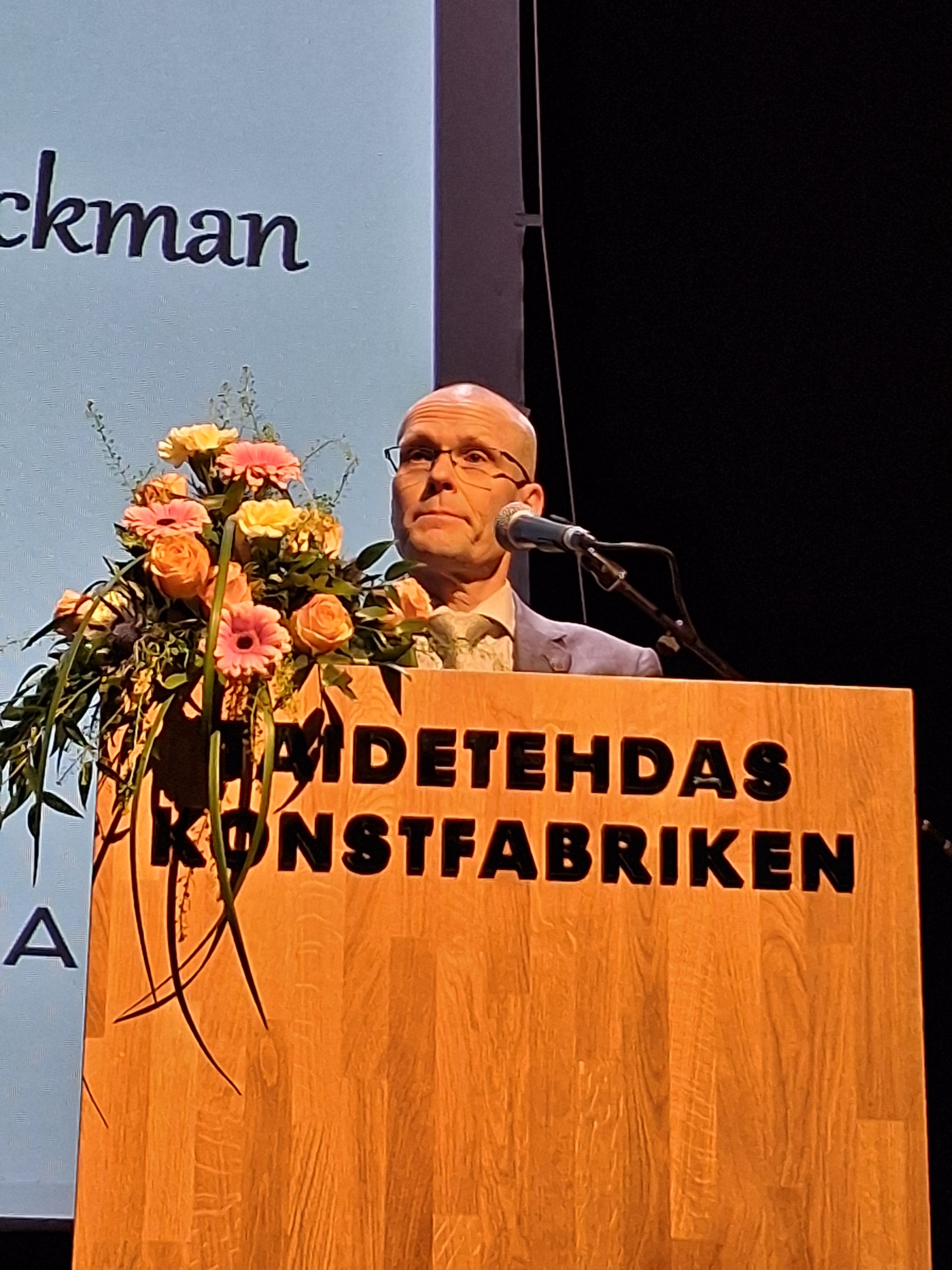 Silmälasipäinen mies vieressään kukkakimppu ja mikrofoni.