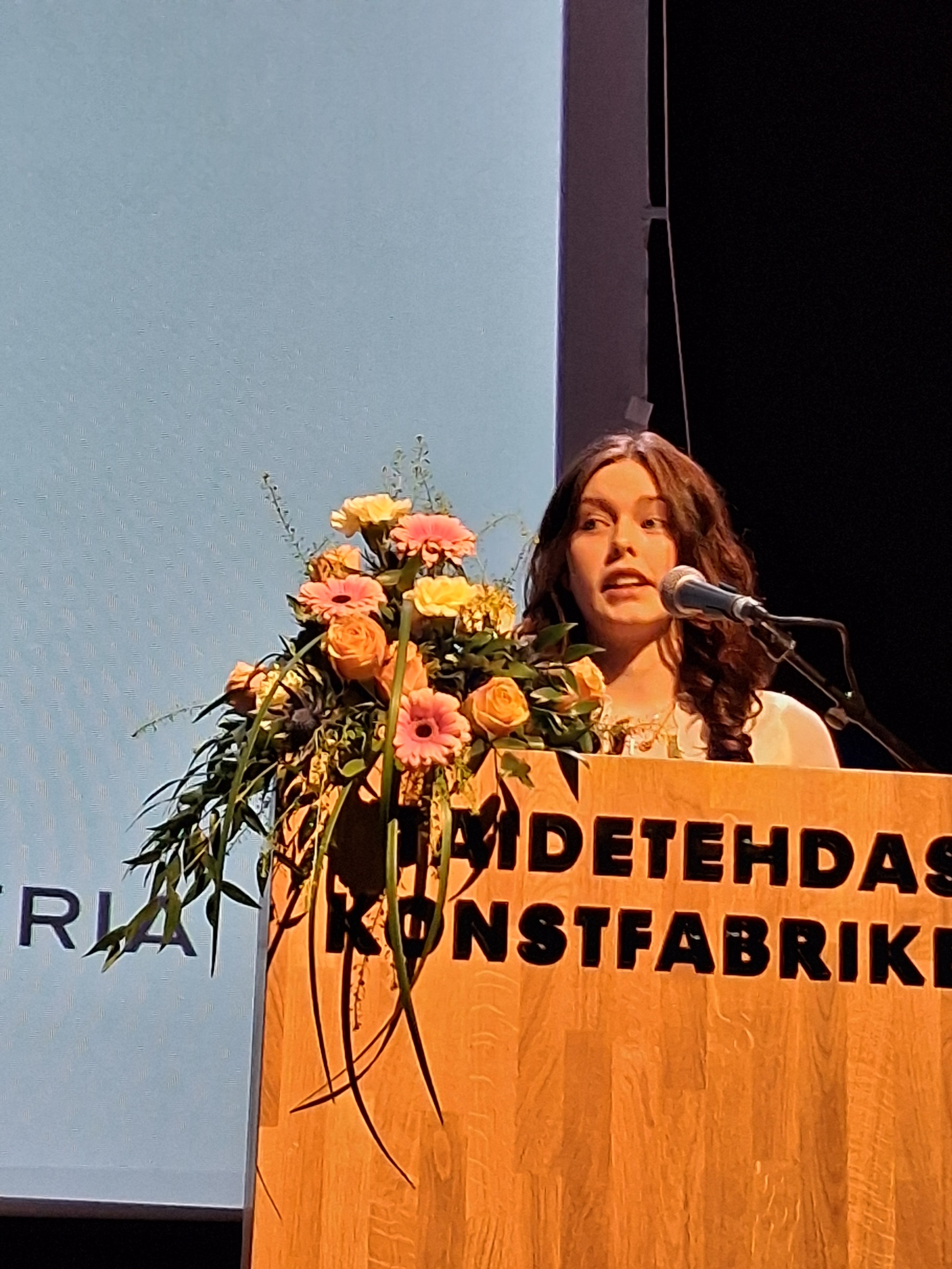 Nuori nainen pitämässä puhetta puhujakorokkeella. Vieressä kukkakimppu ja mikrofoni.