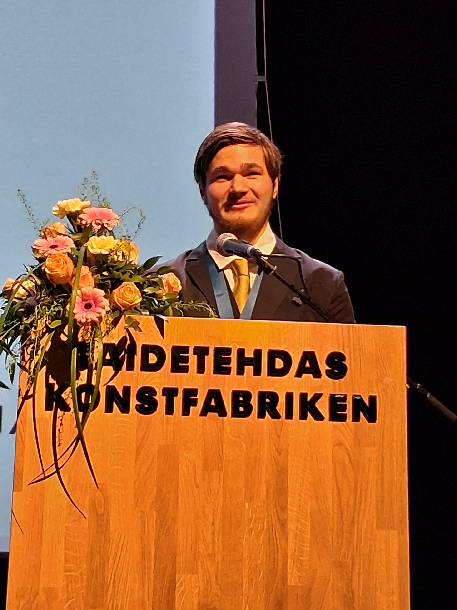 Hymyilevä nuori mies pitämässä puhetta puhujakorokkeella. Vieressä kukkakimppu.