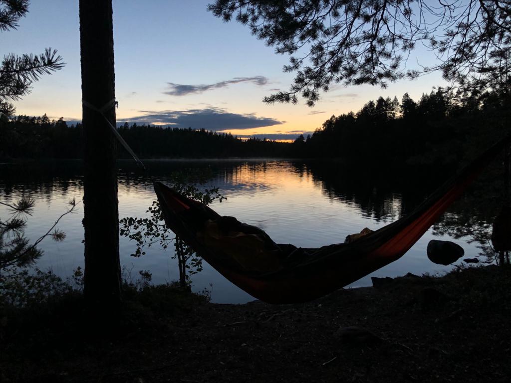 Ihminen makaamassa riippukeinussa hämyistä järvimaisemaa vasten.