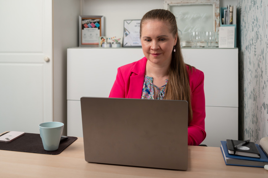 Pinkkiin bleiseriin pukeutunut Anna-Leena Hakuli työskentelee kannettavalla tietokoneella työpöydän ääressä.